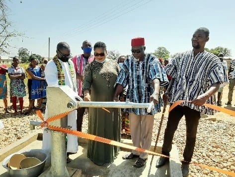 Social Borehole Commission Photo showing Chief of the community in red hat and smock with a female rep from the NGANE-USA cutting the tape as Assembly Member and a Priest look on